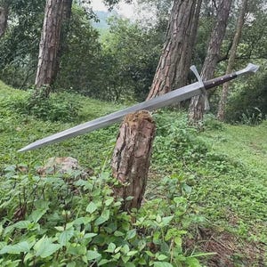 Puede incluir: Una espada larga y plateada con empuñadura y guarda marrón descansa sobre un tocón de árbol. La hoja de la espada es larga y puntiaguda, y la empuñadura es de color marrón oscuro. La guarda es plateada. La espada se exhibe al aire libre, rodeada de vegetación.