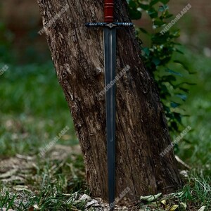 May include: A long, dark sword with a red handle and a black crossguard is leaning against a tree. The blade is a dark gray color, and the handle appears to be made of wood. The sword is displayed outdoors, with the text "THEIRONBLADES" visible.