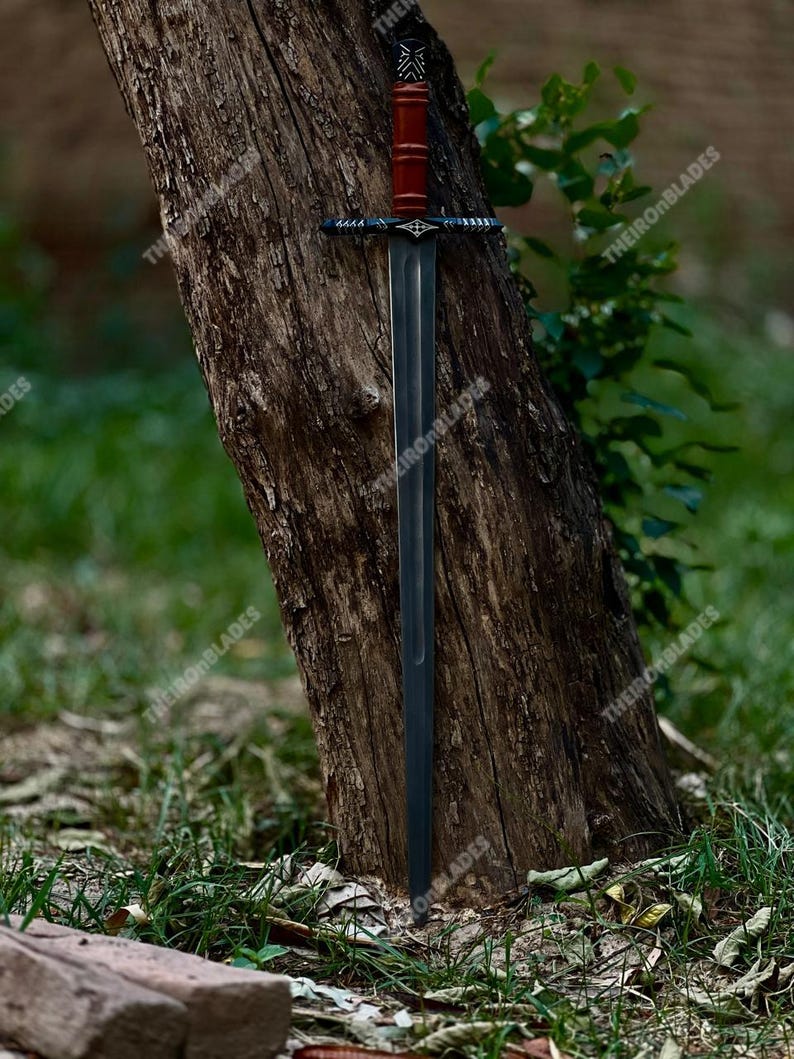 May include: A long, dark sword with a red handle and a black crossguard is leaning against a tree. The blade is a dark gray color, and the handle appears to be made of wood. The sword is displayed outdoors, with the text "THEIRONBLADES" visible.