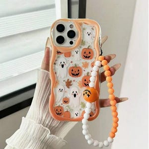 May include: A clear phone case with a wavy orange border featuring a pattern of white ghosts, orange pumpkins, and brown leaves. A beaded wrist strap is attached, consisting of alternating white and orange beads with a pumpkin charm.