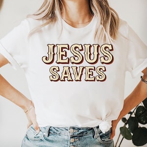 Op de afbeelding: Wit T-shirt met de tekst "JESUS SAVES" in een retro lettertype. De tekst is lichtbruin met een donkerbruine omlijning. Het shirt heeft een klassieke ronde hals.