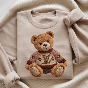 May include: A beige sweatshirt featuring a detailed, embroidered teddy bear. The bear is brown with a sweater in shades of brown and burgundy, and a logo on the front. The sweatshirt has a crew neck and long sleeves.