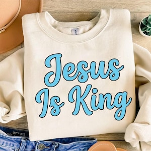 Pode incluir: Camisola creme com a frase "Jesus Is King" em escrita azul claro. O texto tem uma contorno preto. A camisola é de gola redonda e parece ser feita de um material macio.