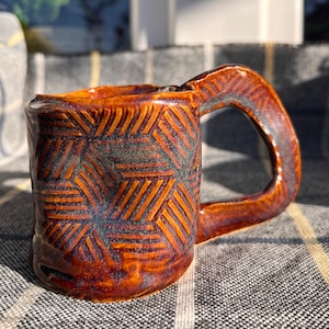 Op de afbeelding: Handgemaakte keramische mok met een geometrisch patroon. De mok is geglazuurd in bruin- en oranjetinten, met vleugjes blauw. De handgreep is dik en afgerond. De mok staat op een geruite stof.