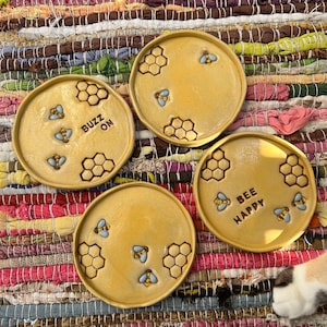 Könnte beinhalten: Vier runde gelbe Keramikschalen mit geprägten Wabenmustern und kleinen blauen und gelben Bienenfiguren. Zwei Schalen enthalten Text, wobei eine BUZZ ON und die andere BEE HAPPY zeigt, angeordnet auf einer strukturierten Oberfläche.