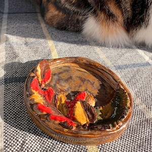 Pode incluir: Um pequeno prato redondo de cerâmica com um esmalte marrom e detalhes de folhas em relevo em vermelho, amarelo e verde. O prato está sobre uma superfície estampada cinza e branca, com parte de um gato calico visível no fundo.