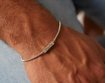 May include: A silver bracelet with a beaded chain design. The bracelet features a rectangular bar engraved with the word "Uncle". The bracelet is worn on a wrist, with the background showing blue denim.