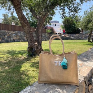 May include: A large, rectangular jute tote bag with sturdy handles. The bag features the letters "TR" in sparkling embellishments and a tassel charm in shades of teal and green. The bag is sitting on a stone wall in a grassy outdoor setting.