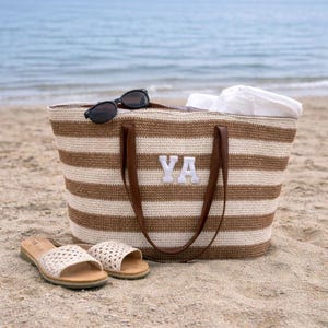 May include: A striped beach tote bag with brown and cream-colored horizontal stripes, leather handles, and the letters "YA" in white. The bag is on a sandy beach with sunglasses and sandals.
