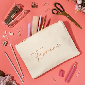Può includere: Un astuccio in tela color crema con il nome "Florence" in oro, circondato da articoli di cancelleria. Include matite, un righello, una pinzatrice, forbici, graffette e un evidenziatore rosa, su sfondo corallo.