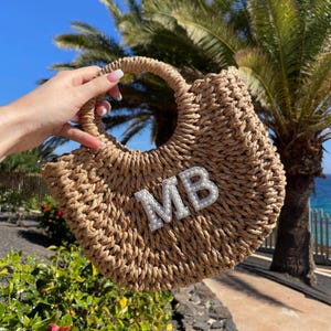 Könnte beinhalten: Beigefarbene Strohhandtasche mit rundem Griff. Die Initialen "MB" sind mit weißen, perlenartigen Perlen besetzt. Die Tasche hat eine geschwungene Form. Im Hintergrund sind Palmen und blauer Himmel zu sehen.
