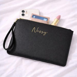 Könnte beinhalten: Schwarze Clutch mit goldenem Reißverschluss und Handschlaufe. Das Wort "Nessy" ist in goldener Schrift auf der Vorderseite geschrieben. Ein Smartphone, ein Portemonnaie mit Kreditkarte und ein Lippenstift sind sichtbar.