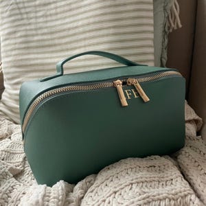 Op de afbeelding: Een teal groene make-uptas met een gouden rits en handvat. De tas heeft de gouden initialen "FE" op de voorkant. De tas ligt op een crèmekleurige gebreide deken, met een gestreept kussen op de achtergrond.