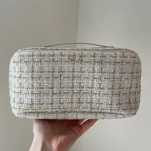 May include: An off-white rectangular cosmetic bag with a woven tweed texture and gold metallic threads. It features a top zip and a small handle. The bag is held by a hand.