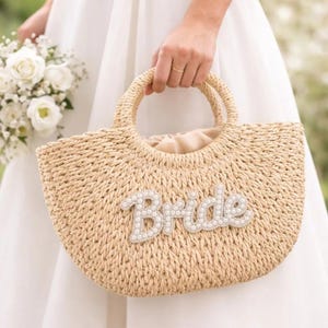 Peut inclure: Sac à main en paille tressée beige avec deux anses circulaires. Le mot "Bride" est épelé en perles blanches sur le devant. L'intérieur du sac est doublé de tissu beige.