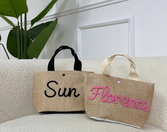 May include: Two woven straw tote bags. One bag has black handles and the word "Sun" embroidered in black. The other bag has cream-colored handles and the word "Florence" embroidered in pink. Both bags have a button closure.