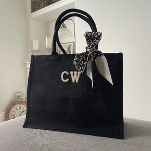 Puede incluir: Bolso tote negro con dos asas y un pañuelo decorativo atado a un asa. El bolso tiene las iniciales "CW" en letras plateadas. El pañuelo es blanco y negro con un diseño de leopardo.