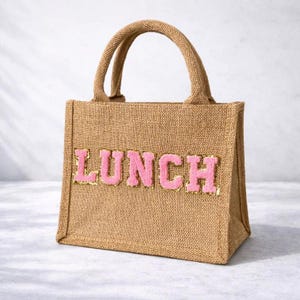 May include: A brown burlap lunch bag with a matching handle. The word "LUNCH" is displayed in pink, fluffy letters with gold trim on the front. The bag is on a white surface. The bag is designed for carrying lunches.