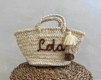 May include: Woven straw tote bag with the name "Lola" embroidered in brown thread. The bag has braided handles and a decorative tassel in shades of beige and brown. The bag is sitting on a woven basket.