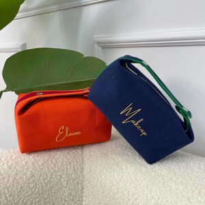 May include: Two fabric cosmetic bags. One is orange with "Elaine" embroidered in gold. The other is navy blue with "Makeup" embroidered in gold. Both have handles and are on a white, textured surface.