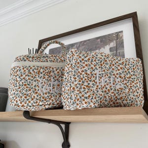 Peut inclure: Deux trousses de maquillage à imprimé floral avec texte brodé blanc. L'une est une petite trousse arrondie avec une poignée, l'autre est une trousse rectangulaire en forme d'oreiller. Les sacs sont sur une étagère en bois.