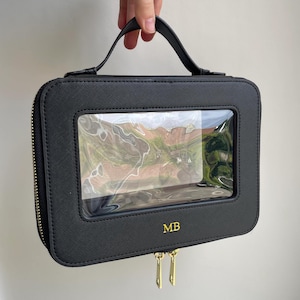 Puede incluir: Estuche de viaje rectangular negro con una ventana transparente y una cremallera dorada. El estuche tiene un asa y las iniciales "MB" en dorado. El estuche está sostenido por una mano.