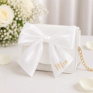 May include: A white leatherette handbag with a large white satin bow. The bag features a gold chain strap and the name "Mia" in gold script. White flowers and petals are in the background.
