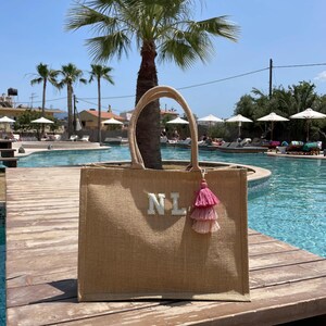 Puede incluir: Un bolso tote de yute color beige con las iniciales "NL" en blanco y una borla rosa. El bolso tiene forma rectangular con un asa. El fondo muestra una piscina y palmeras.