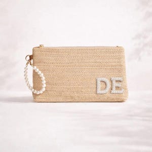 Könnte beinhalten: Beige gewebte Clutch mit den Buchstaben "DE" aus silbernen Strasssteinen. Die Clutch hat einen goldenen Reißverschluss und eine Perlen-Handschlaufe. Die rechteckige Tasche ist aus Stroh-ähnlichem Material.