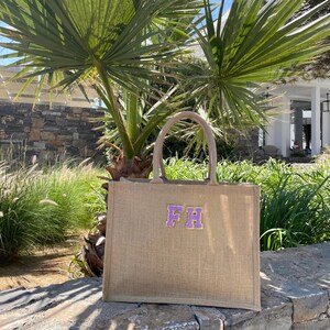 May include: A tan jute tote bag with a pair of purple embroidered initials "FH". The bag has a rectangular shape with a wide opening and two handles. The bag is sitting on a stone wall, with palm trees in the background.