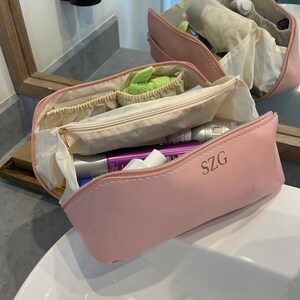 May include: Pale pink cosmetic bag with a zip closure, holding toiletries. The bag has the initials "SZG" printed on the side. Another similar bag is visible in the background, both open and filled with items.
