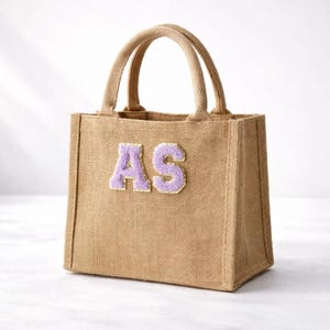 Könnte beinhalten: Eine rechteckige Tragetasche aus Jute mit zwei Griffen. Die Tasche hat die gestickten Buchstaben "AS" in einem hellen Lila. Die Tasche hat eine natürliche beige Farbe und scheint leer zu sein.