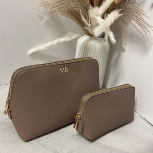 May include: Two taupe cosmetic bags with gold zips. The larger bag has the gold initials "MB". The bags are different sizes and are on a light surface. A white vase with dried flowers is in the background.