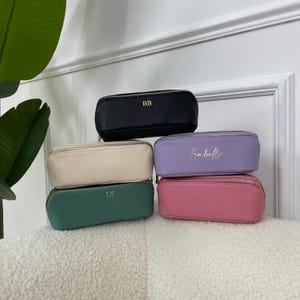 May include: Five rectangular pencil cases in a stack, in black, cream, lavender, pink, and teal. Each case has a gold zip and gold lettering. The black case has "BB", the lavender case has "Isabell", and the teal case has "LS".