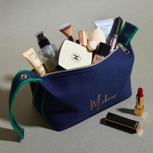 May include: A navy blue makeup bag with a green trim, filled with various cosmetic products, including brushes, tubes, and compacts. The bag has the word "Makeup" in gold script. A red lipstick and a black and gold lipstick case are next to the bag.