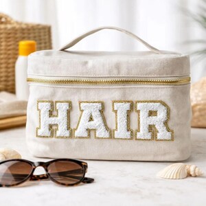 May include: A beige cosmetic bag with a gold zipper and a handle. The word "HAIR" is embroidered in white, fluffy letters with gold trim. Tortoiseshell sunglasses and seashells are in the foreground.