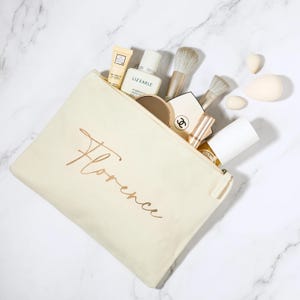 Può includere: Pochette per cosmetici in tela color crema con il nome "Florence" in oro. La borsa aperta rivela pennelli per il trucco, ciprie, rossetti e prodotti per la cura della pelle. Sfondo in marmo bianco.