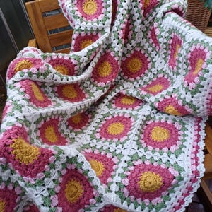 Peut inclure: Une couverture au crochet faite à la main avec un motif floral. La couverture présente un motif répété de fleurs roses, jaunes et vertes sur un fond blanc. Elle est drapée sur une chaise en bois, mettant en valeur sa texture et ses couleurs.