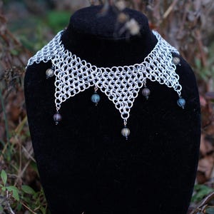 May include: A silver chainmail necklace with a scalloped edge and dangling beads. The necklace is displayed on a black mannequin against a blurred outdoor background. The beads are a mix of colours.