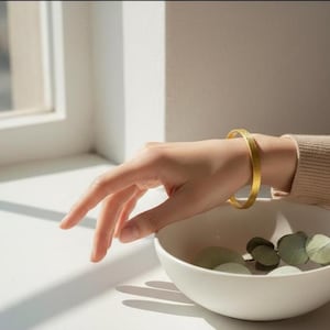 Peut inclure: Un bracelet jonc doré avec une surface texturée est porté au poignet. La main tend la main vers un bol blanc contenant des éléments décoratifs verts. L'arrière-plan comprend une fenêtre et une surface blanche.