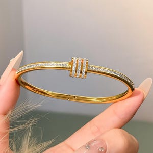 Può includere: Un bracciale rigido dorato con un elemento decorativo centrale e pietre chiare scintillanti. Il bracciale ha una chiusura a cerniera ed è tenuto da una mano con unghie lunghe e curate. È un gioiello.