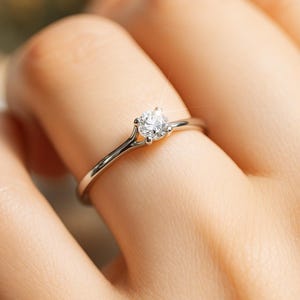 Peut inclure: Gros plan d'une bague en argent avec une pierre précieuse claire et ronde. La bague est portée au doigt, et le sertissage est délicat. La pierre précieuse scintille, captant la lumière.