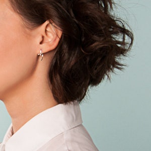 May include: A woman with short, dark brown, wavy hair styled in a side part. She is wearing a white collared shirt with a sleeveless design. The background is a light blue.