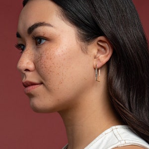 Könnte beinhalten: Eine Frau mit langen dunklen Haaren und Sommersprossen schaut zur Seite. Sie trägt ein weißes Tanktop und einen kleinen Creolenohrring am linken Ohr.