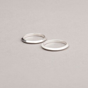 May include: Two silver rings with a flat top and angled sides are displayed on a light gray surface. The rings are simple in design and appear to be made of metal. The rings are circular and have a polished finish.