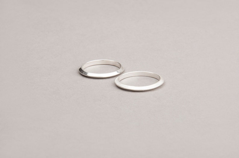 May include: Two silver rings with a flat top and angled sides are displayed on a light gray surface. The rings are simple in design and appear to be made of metal. The rings are circular and have a polished finish.