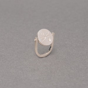 May include: A silver ring with a spinning disc featuring a design of a bird and an arrow. The ring is open and has a smooth, polished finish.
