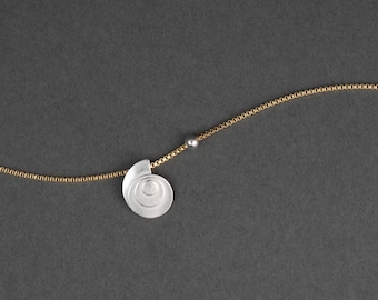 Puede incluir: Collar con colgante plateado en forma de espiral. La fina cadena dorada tiene una pequeña cuenta gris. La joya se muestra sobre un fondo gris oscuro.