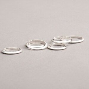 May include: Five silver rings with a minimalist design are arranged on a gray surface. The rings have a smooth, polished finish and a simple, rounded shape. The rings are a jewelry item.