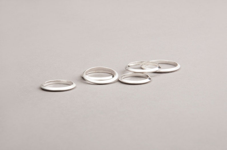 May include: Five silver rings with a minimalist design are arranged on a gray surface. The rings have a smooth, polished finish and a simple, rounded shape. The rings are a jewelry item.